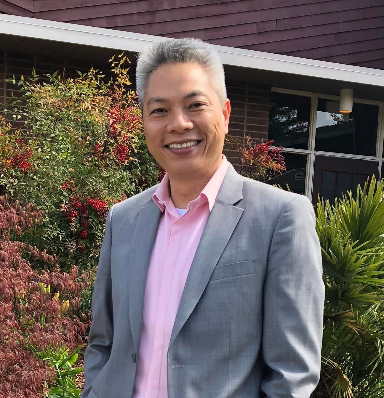 A person with short, grey hair wearing a light pink shirt and grey blazer, smiling outdoors with foliage in the background.