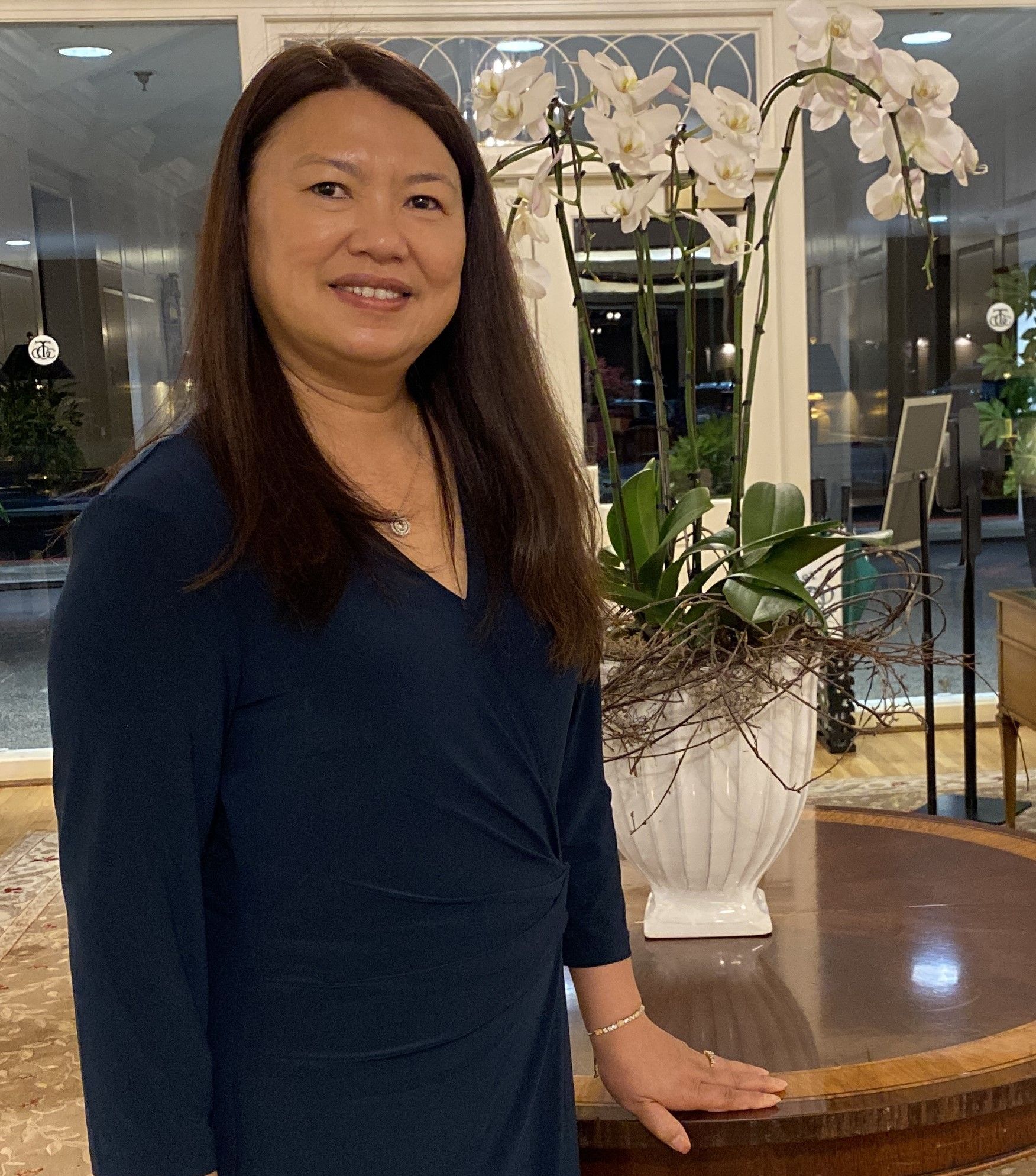 A person with long brown hair in a navy blue dress standing next to a white planter with white orchids in a bright room.