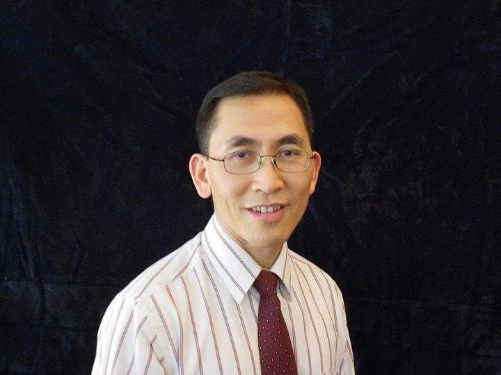 A professional headshot of a person wearing glasses, a striped white shirt, and a dark red tie against a dark background.