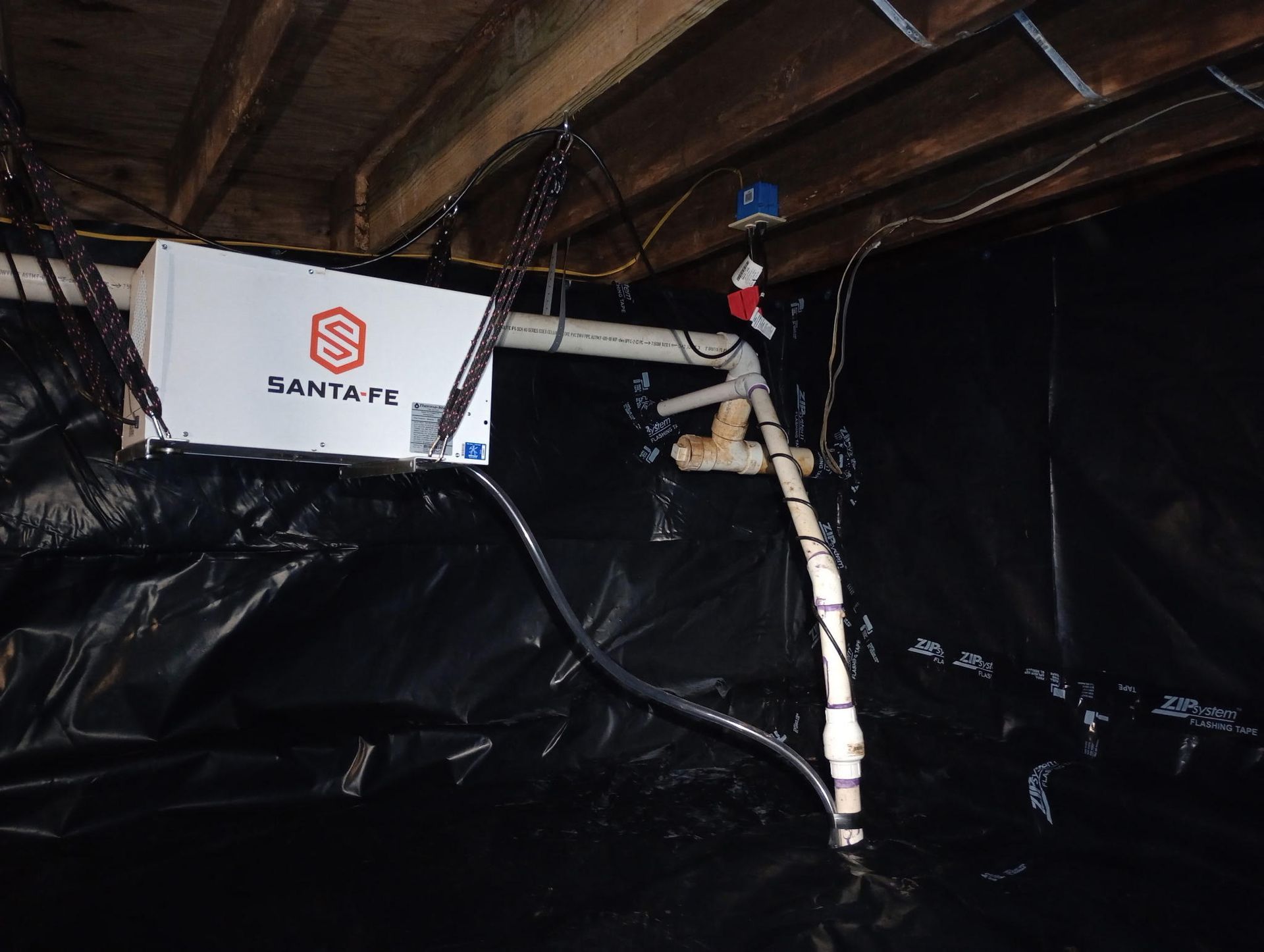 A Santa Fe crawl space dehumidifier suspended from wooden joists above a dark, plastic-covered ground surface.