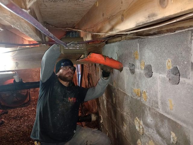 A person in a crawl space installs a piece of orange flashing near a wood floor joist and a concrete block wall.