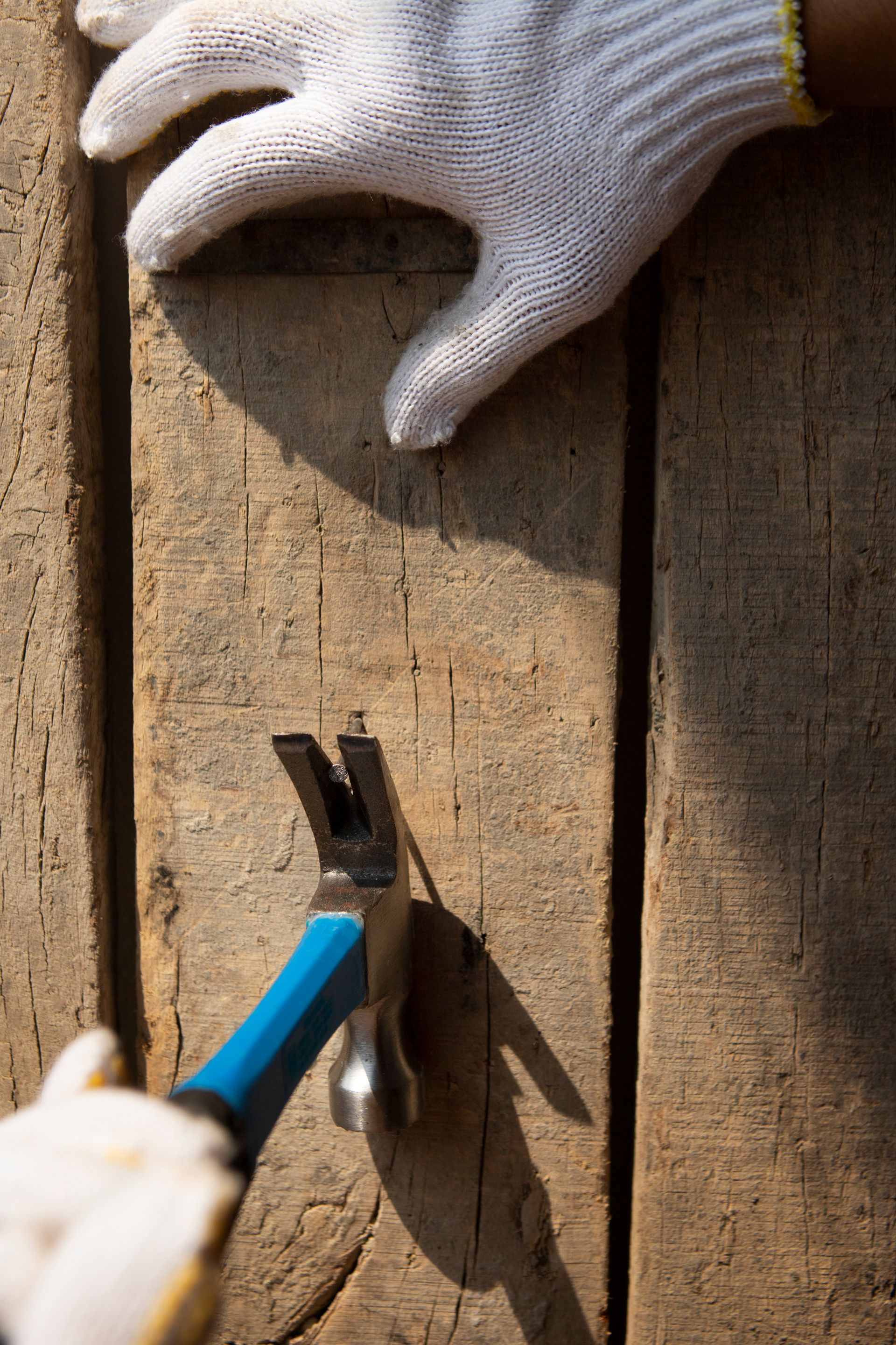 Gloved hands using a hammer to remove a nail from a weathered wooden surface.