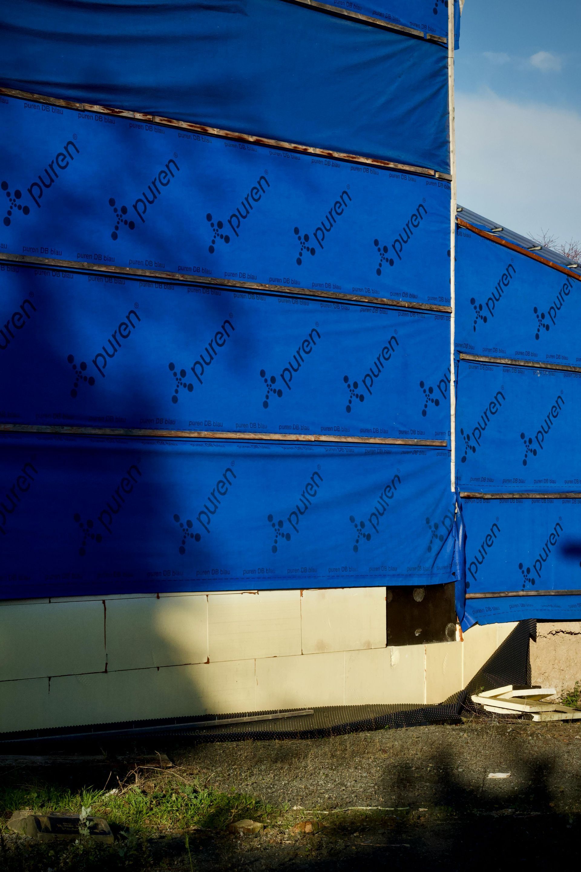 Blue protective house wrap covering the exterior walls of a building under construction, above an unfinished foundation.