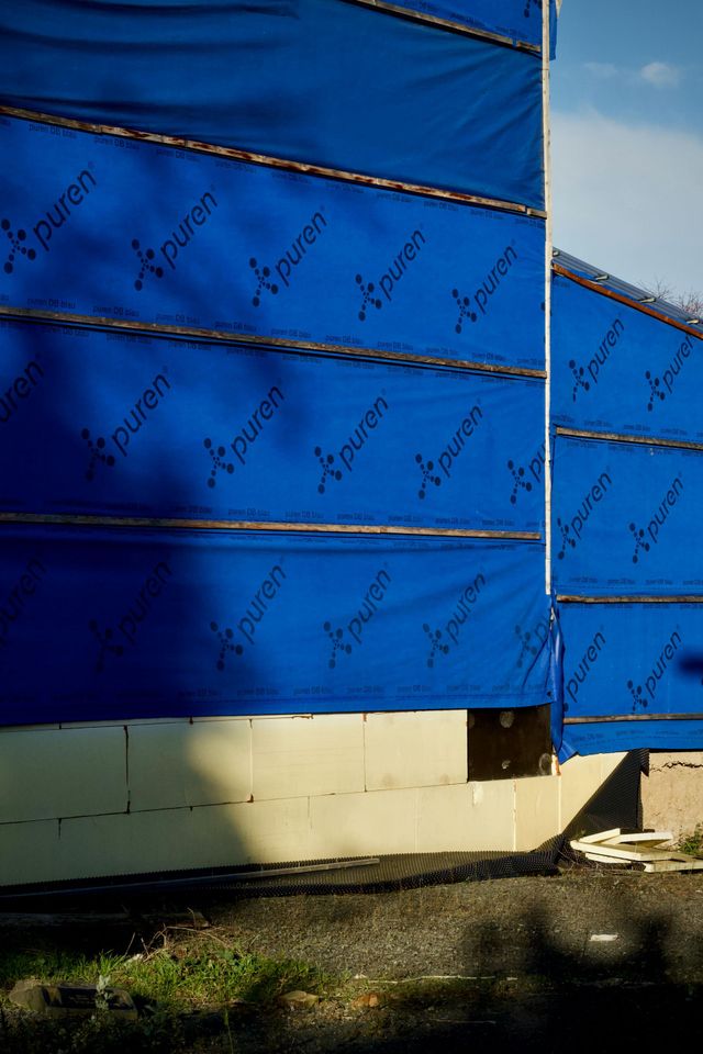 Blue protective house wrap covering the exterior walls of a building under construction, above an unfinished foundation.