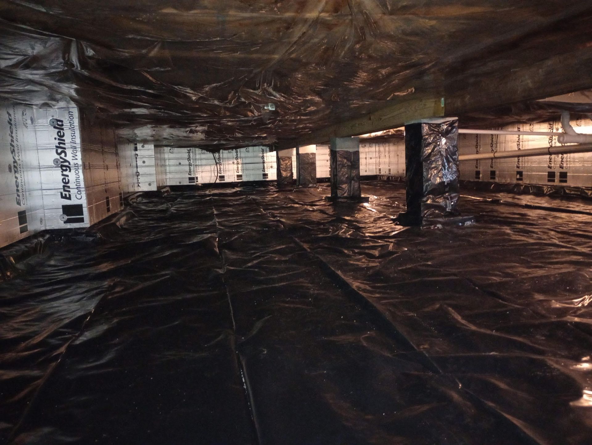 A crawl space with insulated walls, a black plastic vapor barrier on the floor, and support pillars.