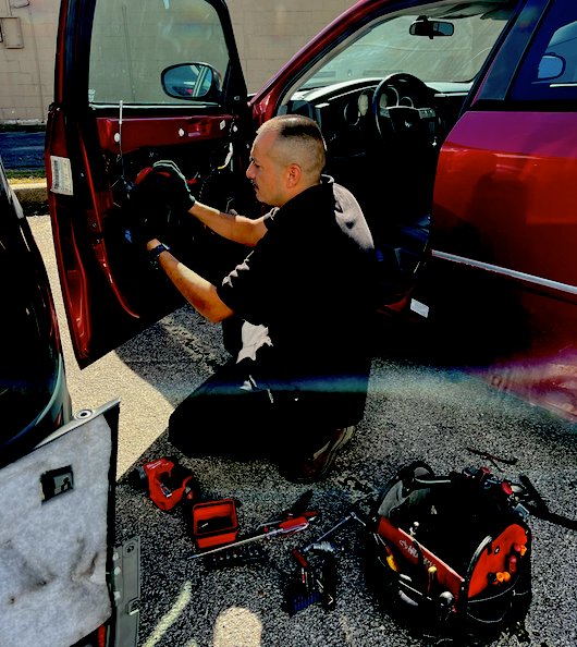 Mechanic working on car door with tools on the ground outside. Car is red.