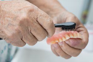 Hands brushing a pink denture with a black toothbrush; close-up.