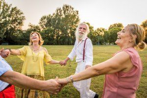 Group of older adults holding hands, laughing, and dancing in a circle outdoors on a sunny day.