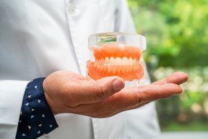 Dentist holds a model of dentures in their open palm, in front of a green background.