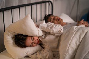 Woman covers ears with pillow, annoyed by snoring man in bed.