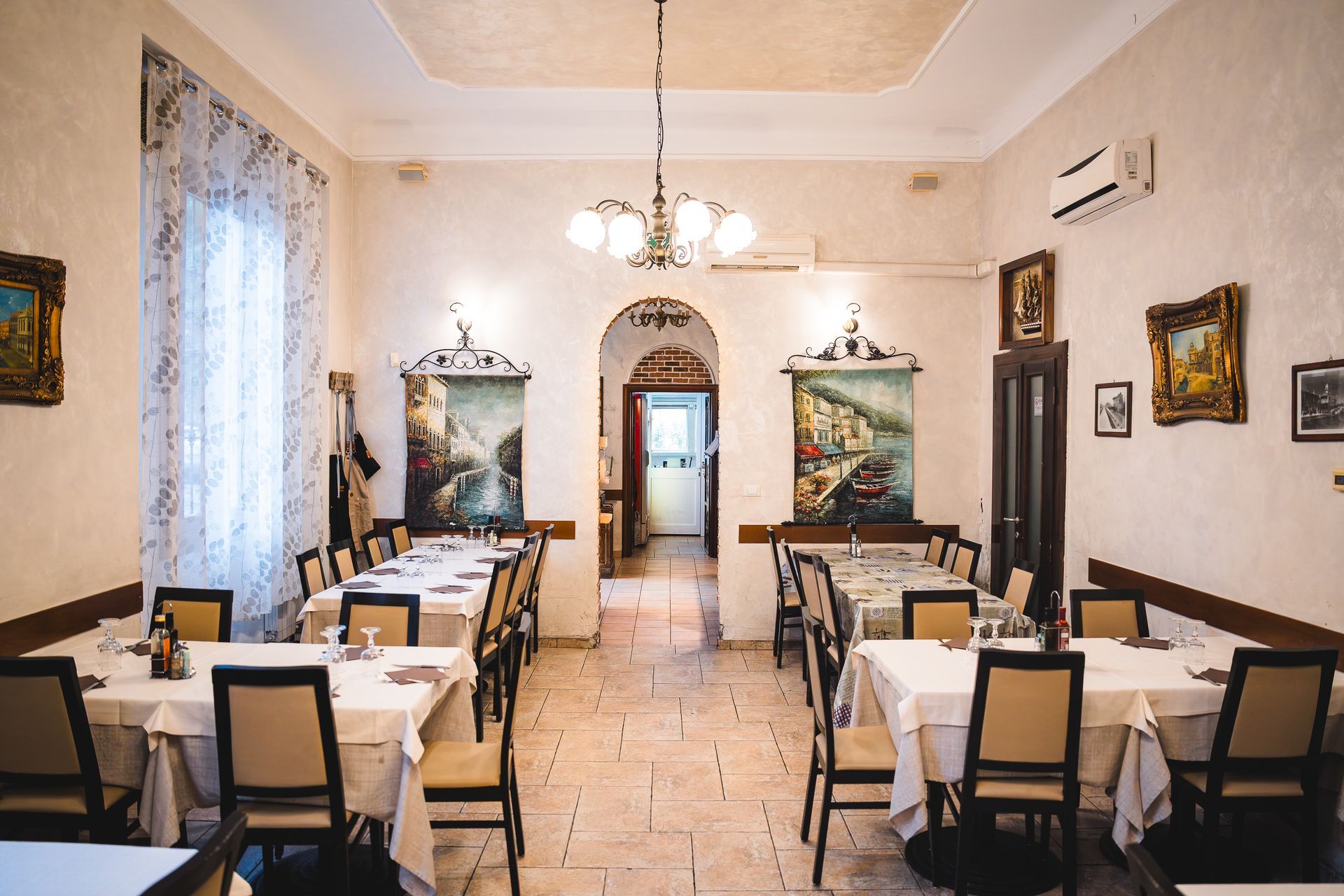 Interno del ristorante: tavoli apparecchiati per cenare, quadri incorniciati alle pareti, porta ad arco che conduce a un'altra sala.