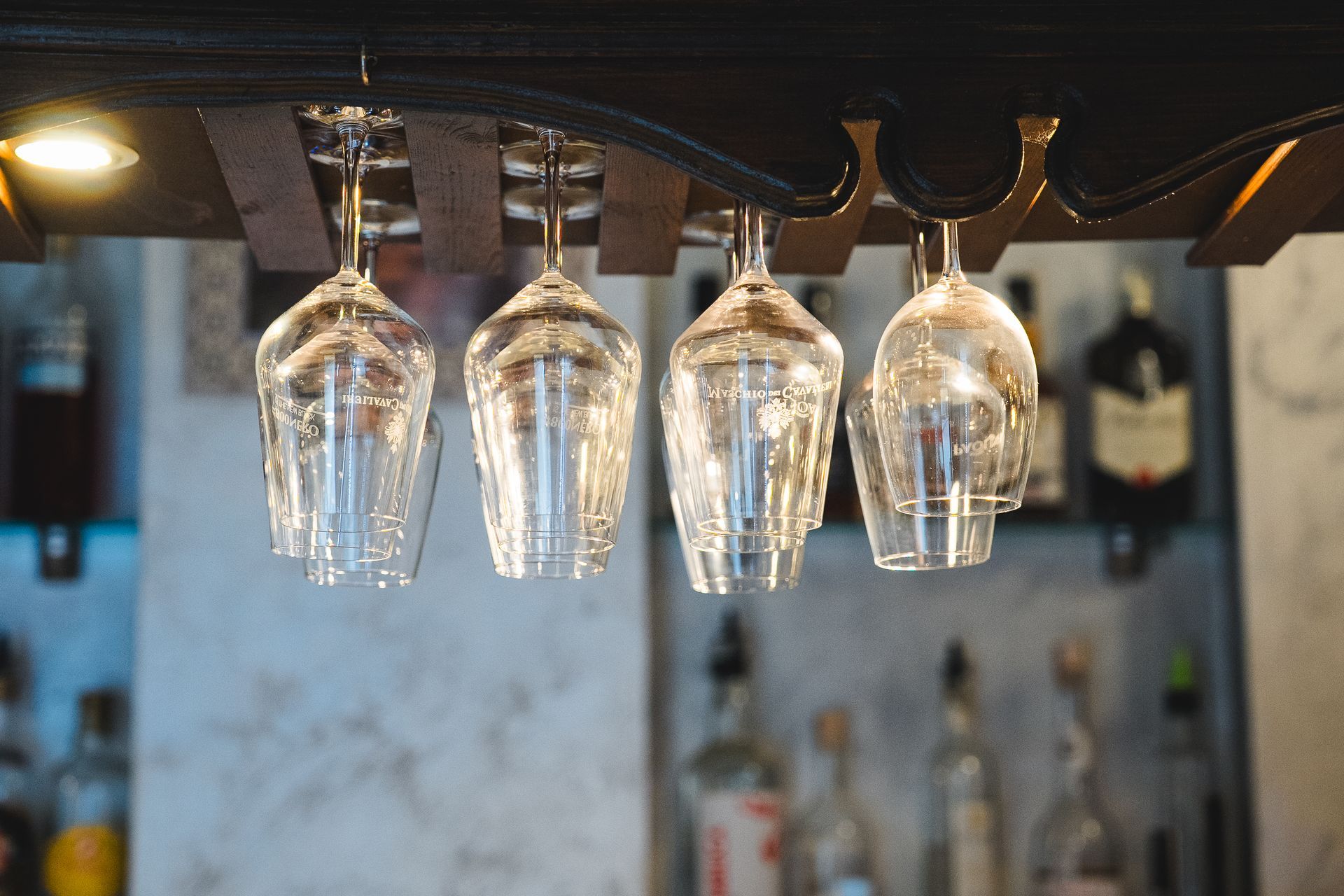 Quattro bicchieri da vino trasparenti con stelo appesi capovolti a un portaoggetti da bar in legno. Sfondo del bar.