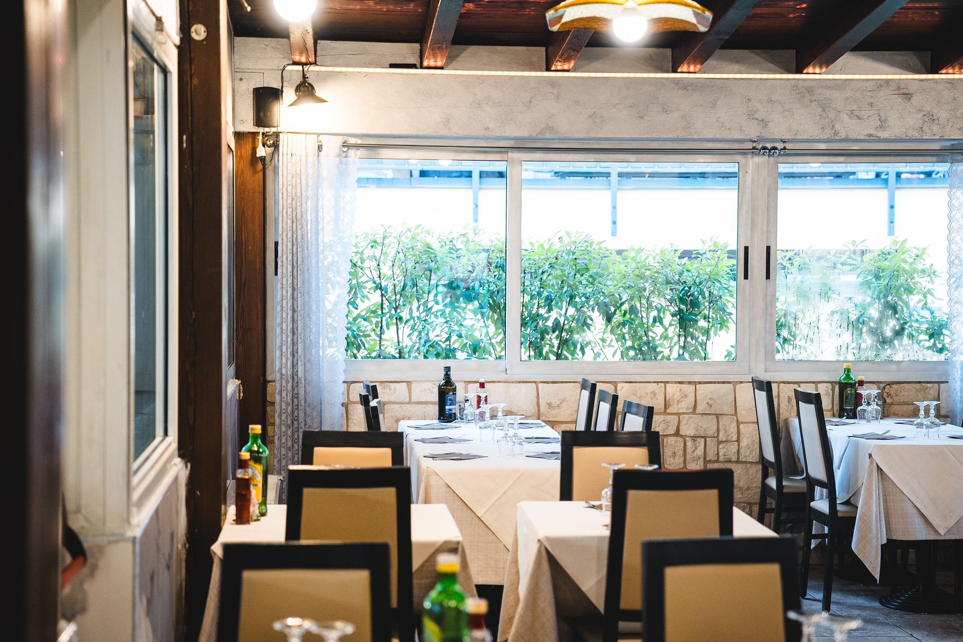 Interno di un ristorante con tavoli apparecchiati per cenare vicino a una finestra con vista sul verde.