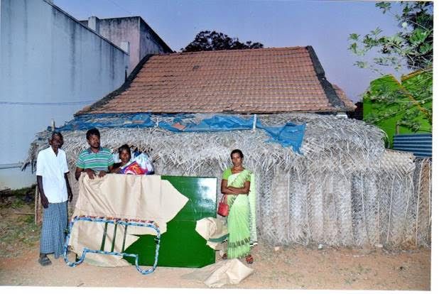 People standing beside a makeshift stall with a thatched roof and green wall outdoors