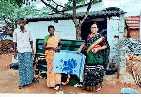 Three people standing beside a blue donation box outside a small house with a tree and stone wall