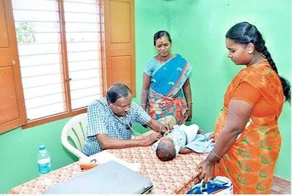 Man seated at a table examining a child’s head while two women stand nearby in a clinic room
