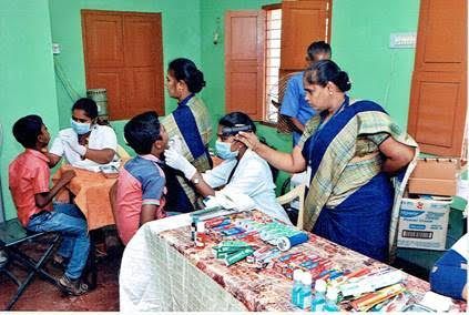 Health workers administering care in a clinic room, with seated patients and supplies on a table.