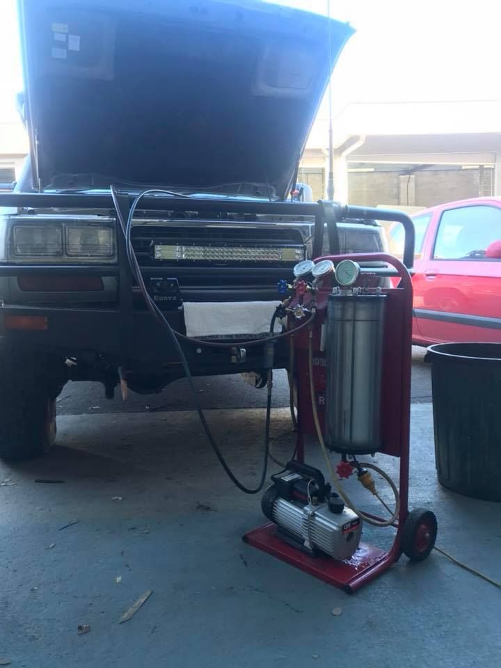 A vehicle with its hood up, connected to an automotive fluid recycling machine on a rolling cart.