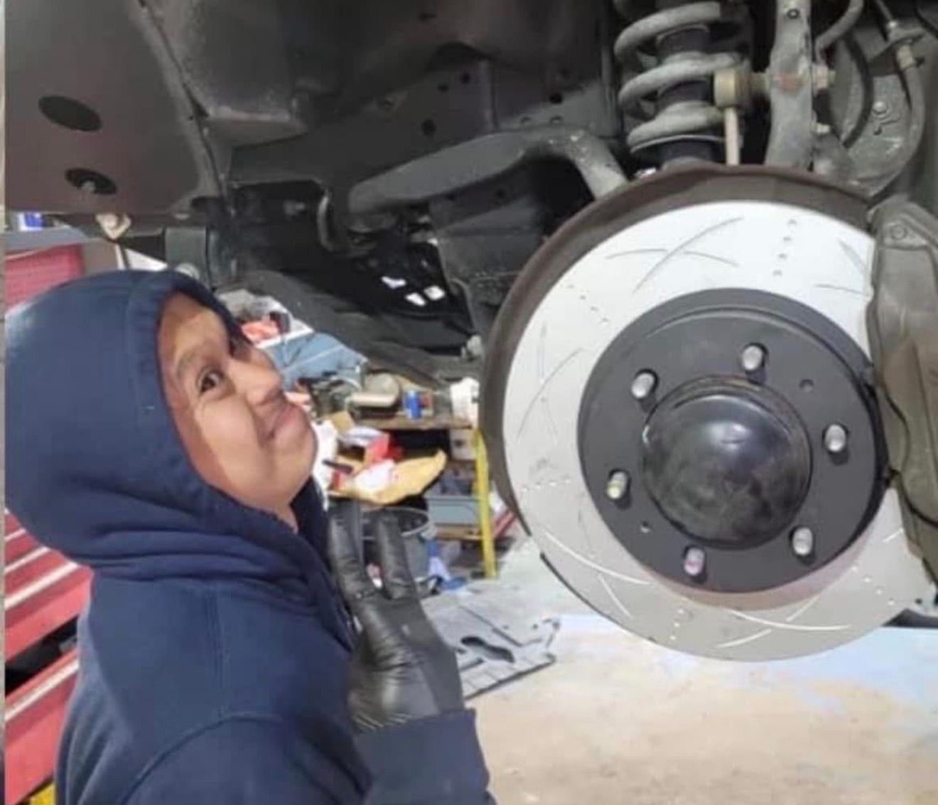 A person in a blue hoodie and black gloves makes a peace sign while standing by a vehicle with an exposed brake rotor.