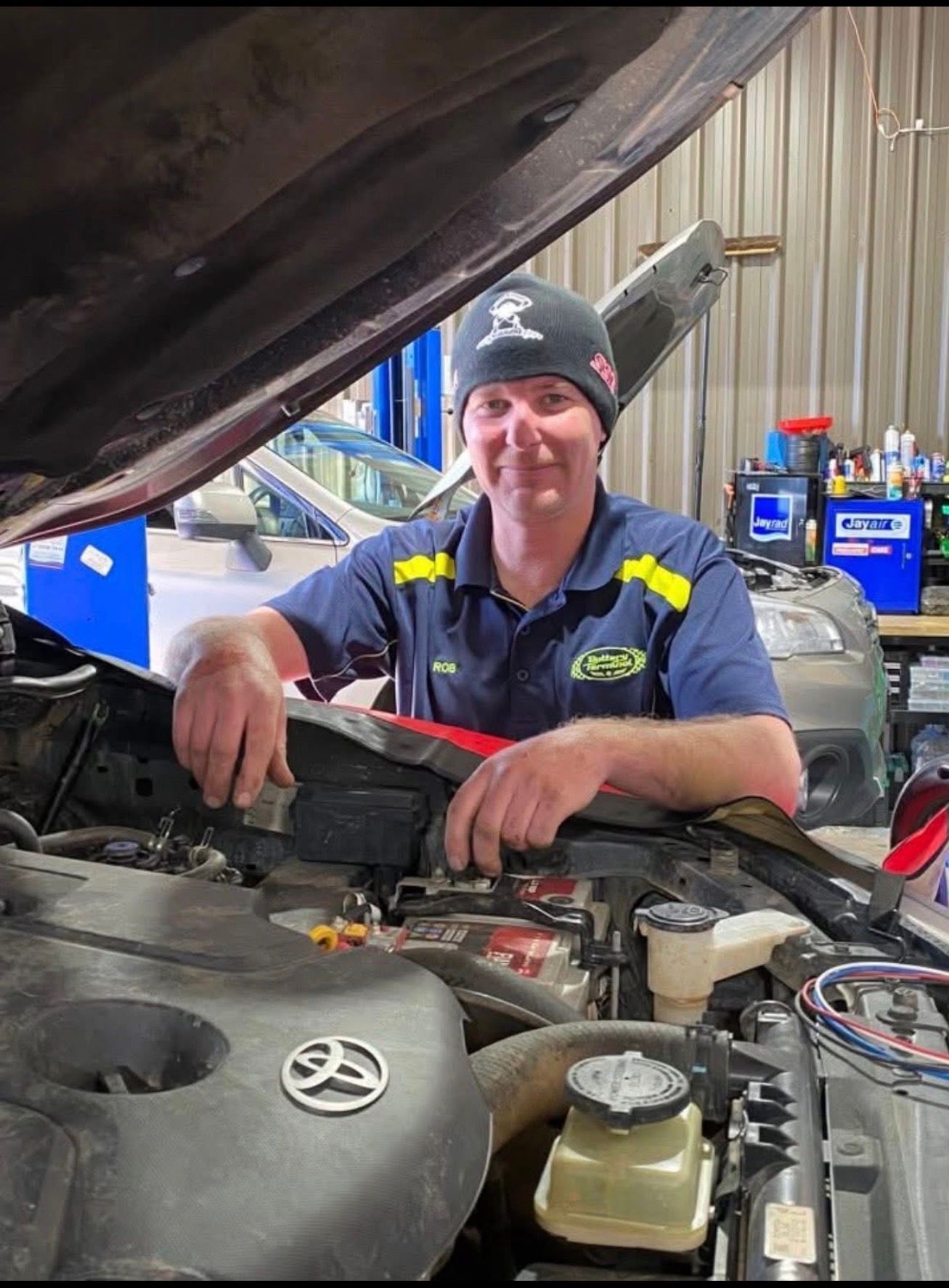A mechanic in a blue uniform and black beanie leans over an open car hood in a repair shop.