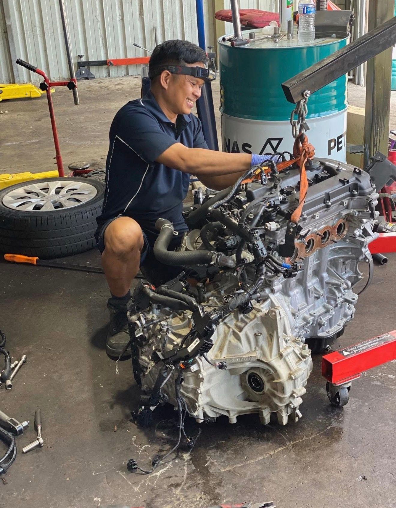 A mechanic in a blue shirt kneels in a garage, using a crane to lift a car engine.