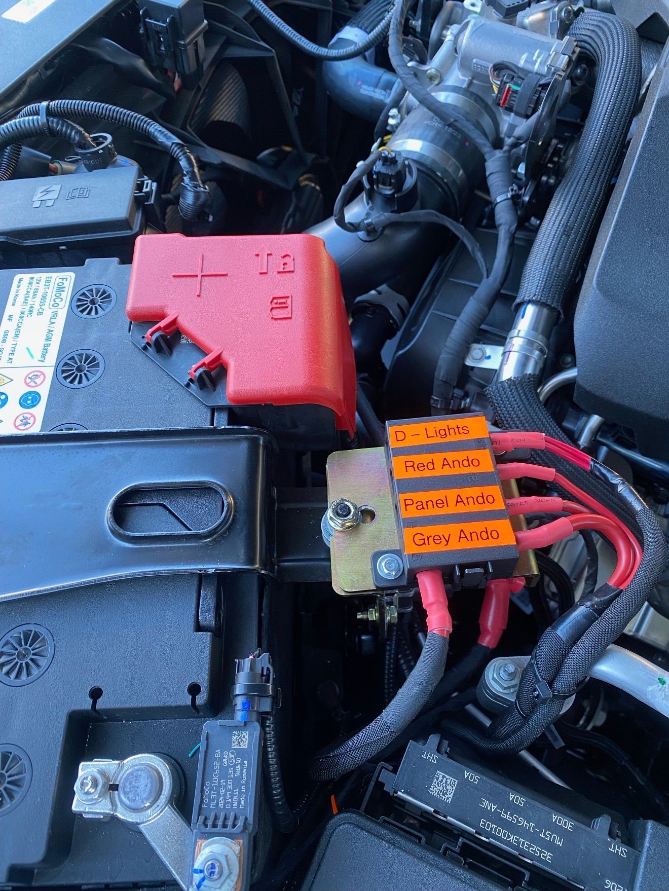 A car battery with a positive terminal red cover and a fuse block with four labeled wires connected to the terminal.