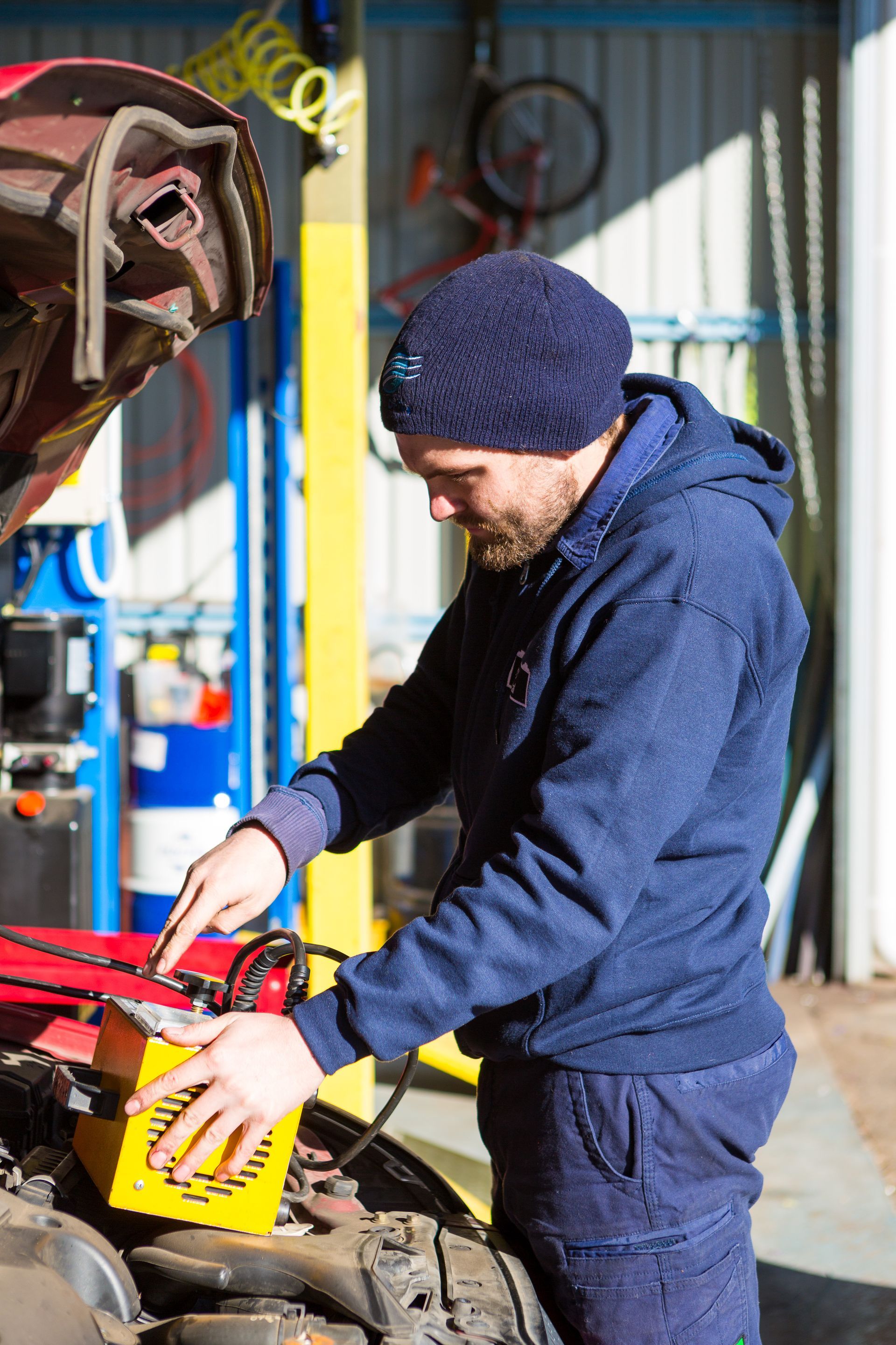 A person in a blue hoodie and beanie works under the hood of a car in a garage, holding a yellow device.