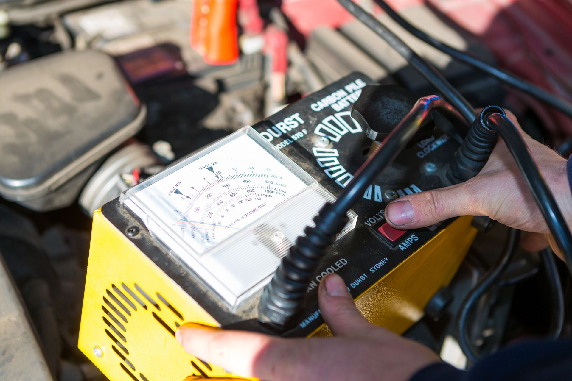 Hands adjusting a yellow battery charger near a car engine, indicating charging.
