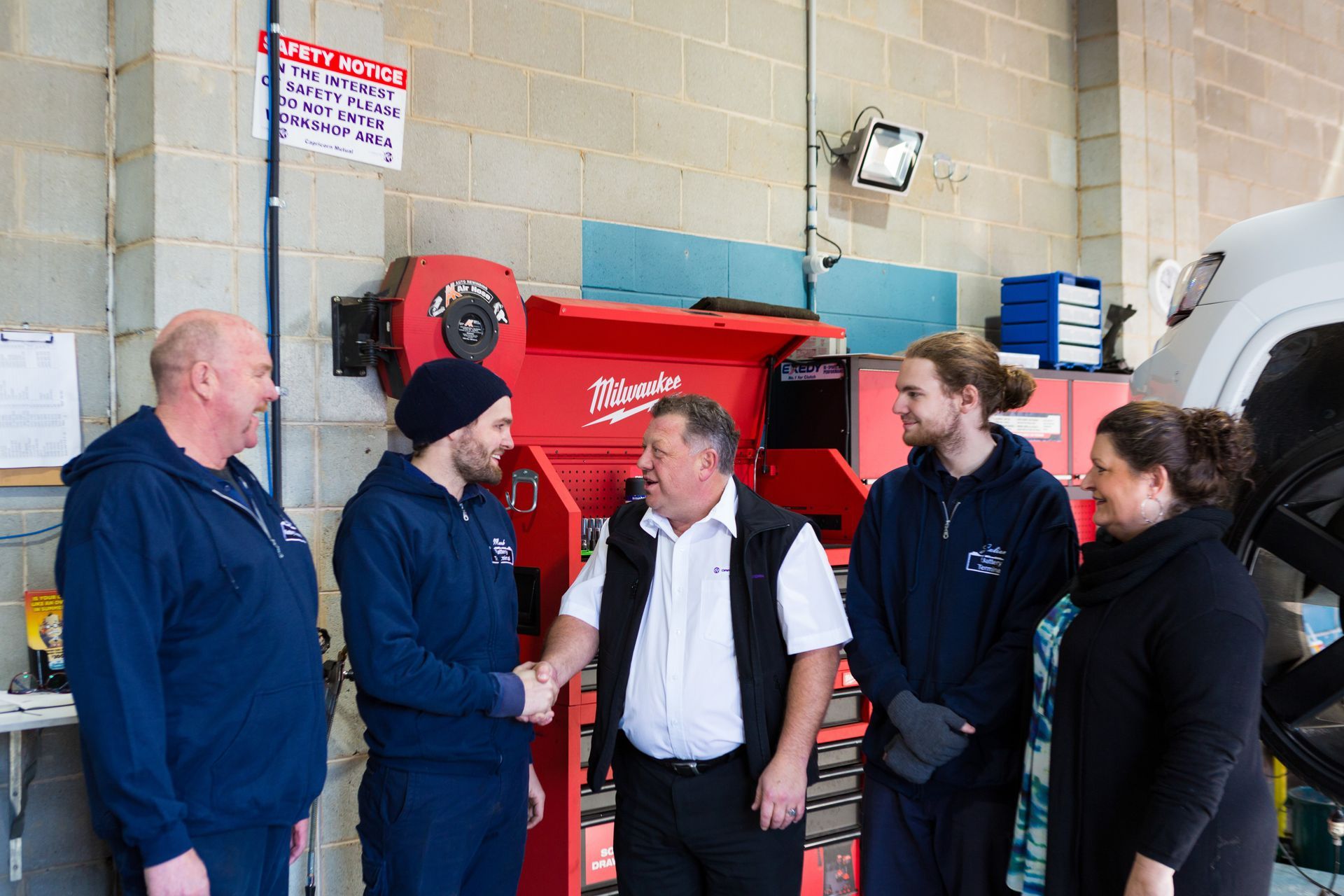 Five people stand in a workshop near a red tool cabinet, with one person shaking hands with another.