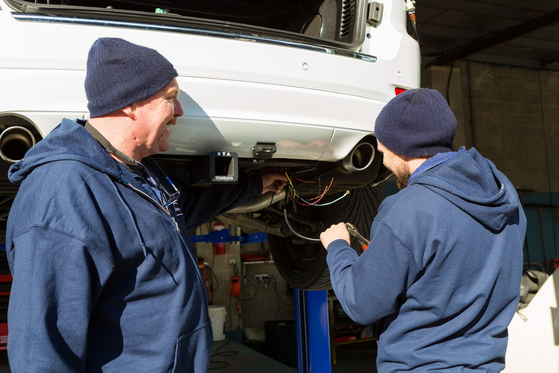 Two mechanics, wearing beanies and blue hoodies, work on the undercarriage of a white SUV in a shop.