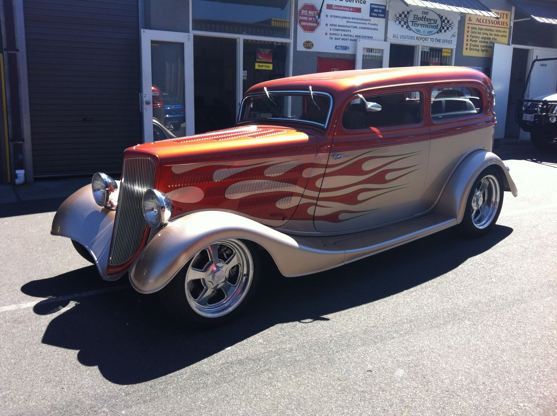 Custom orange and tan hot rod with flames parked in front of a shop.