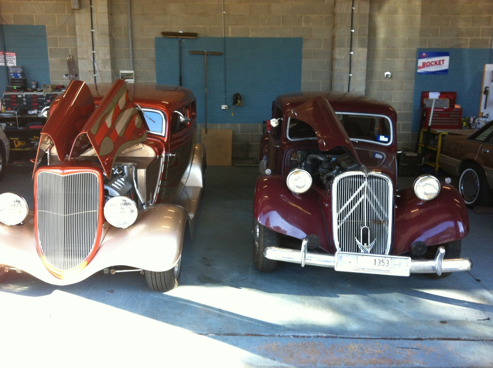 Two classic cars with open hoods, one tan, the other burgundy, parked in a garage.