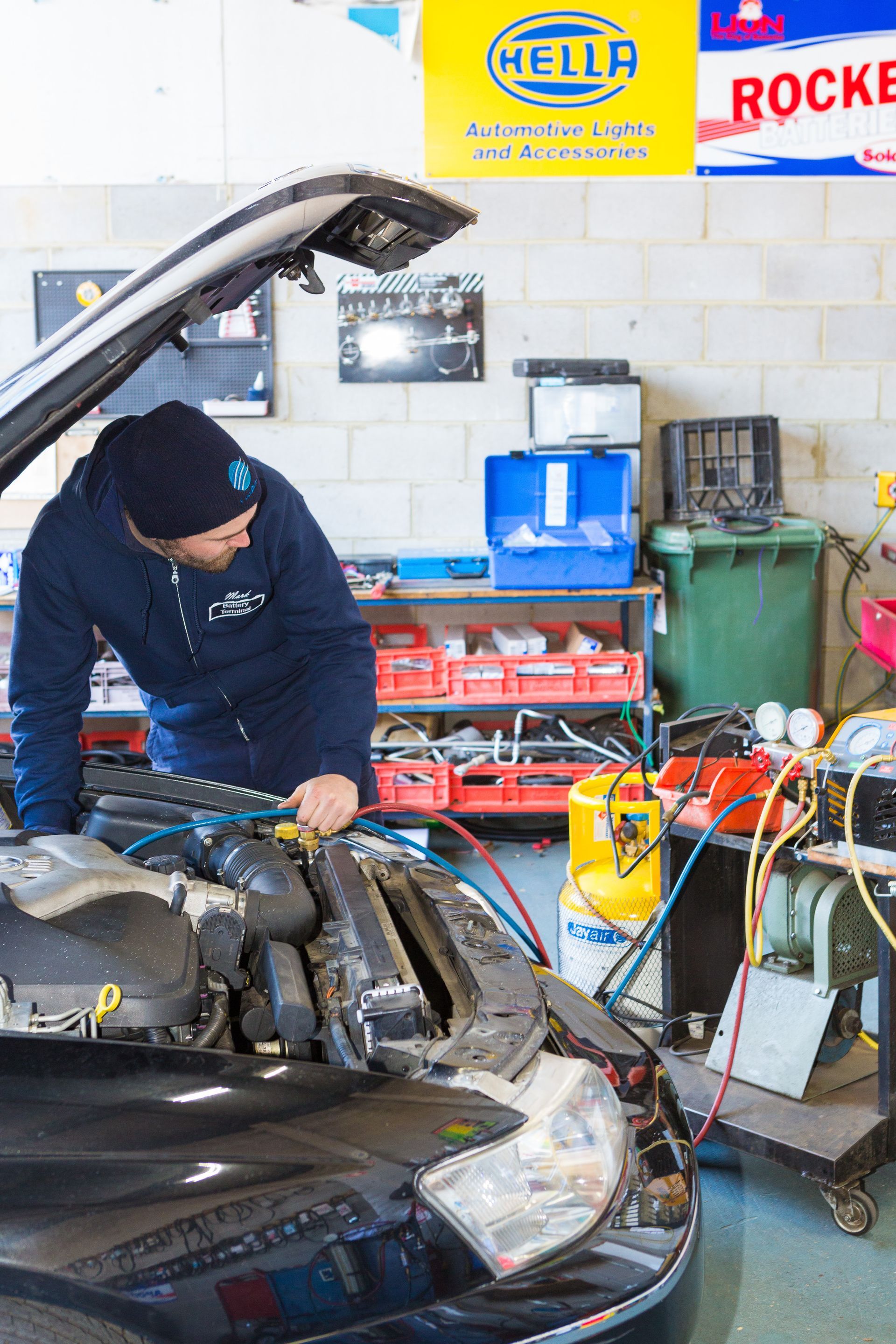 A technician in a blue uniform and beanie works on an open car engine in an auto repair shop with diagnostic equipment.