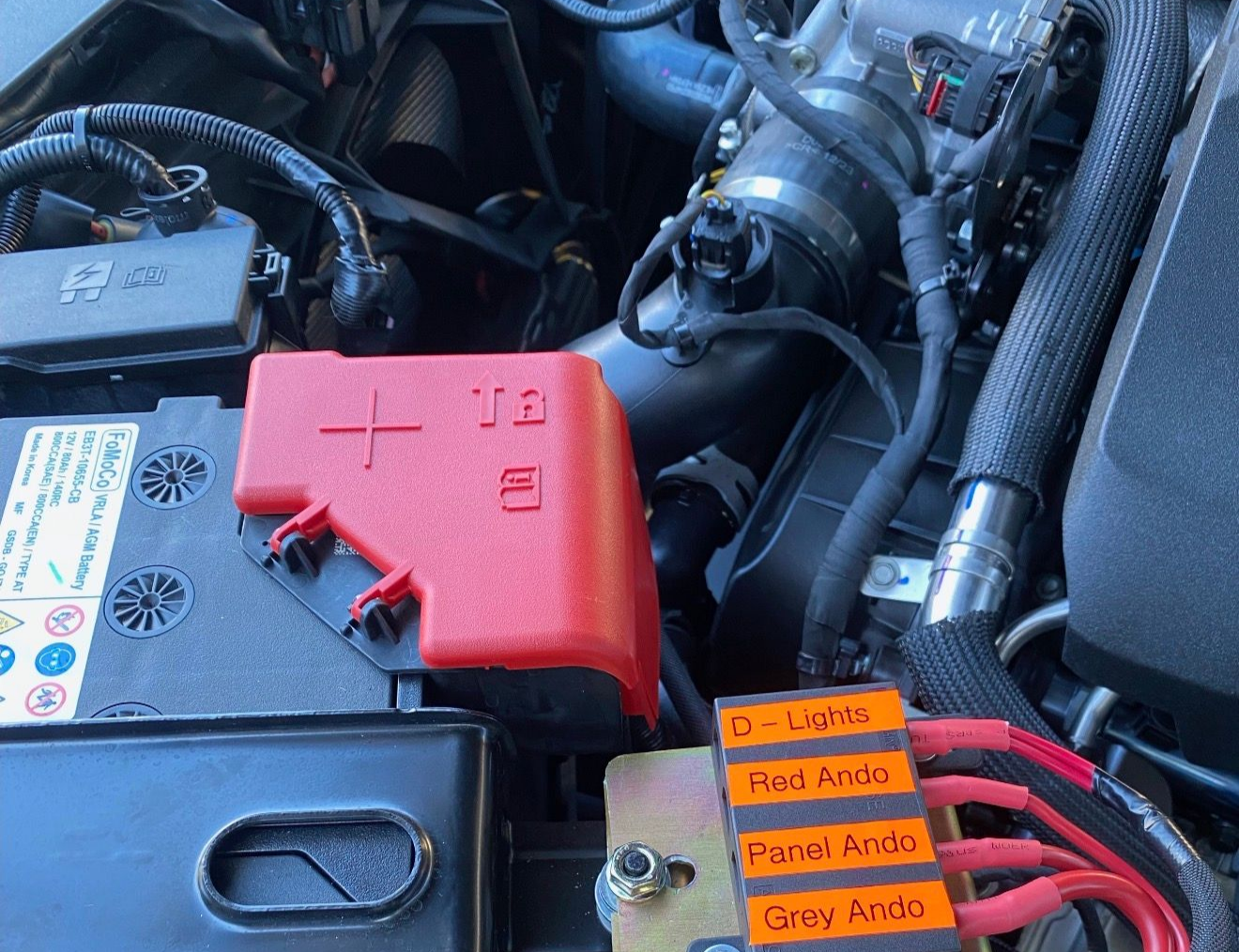 A car battery with a positive terminal cover and an aftermarket fuse block with labeled red wires connected.