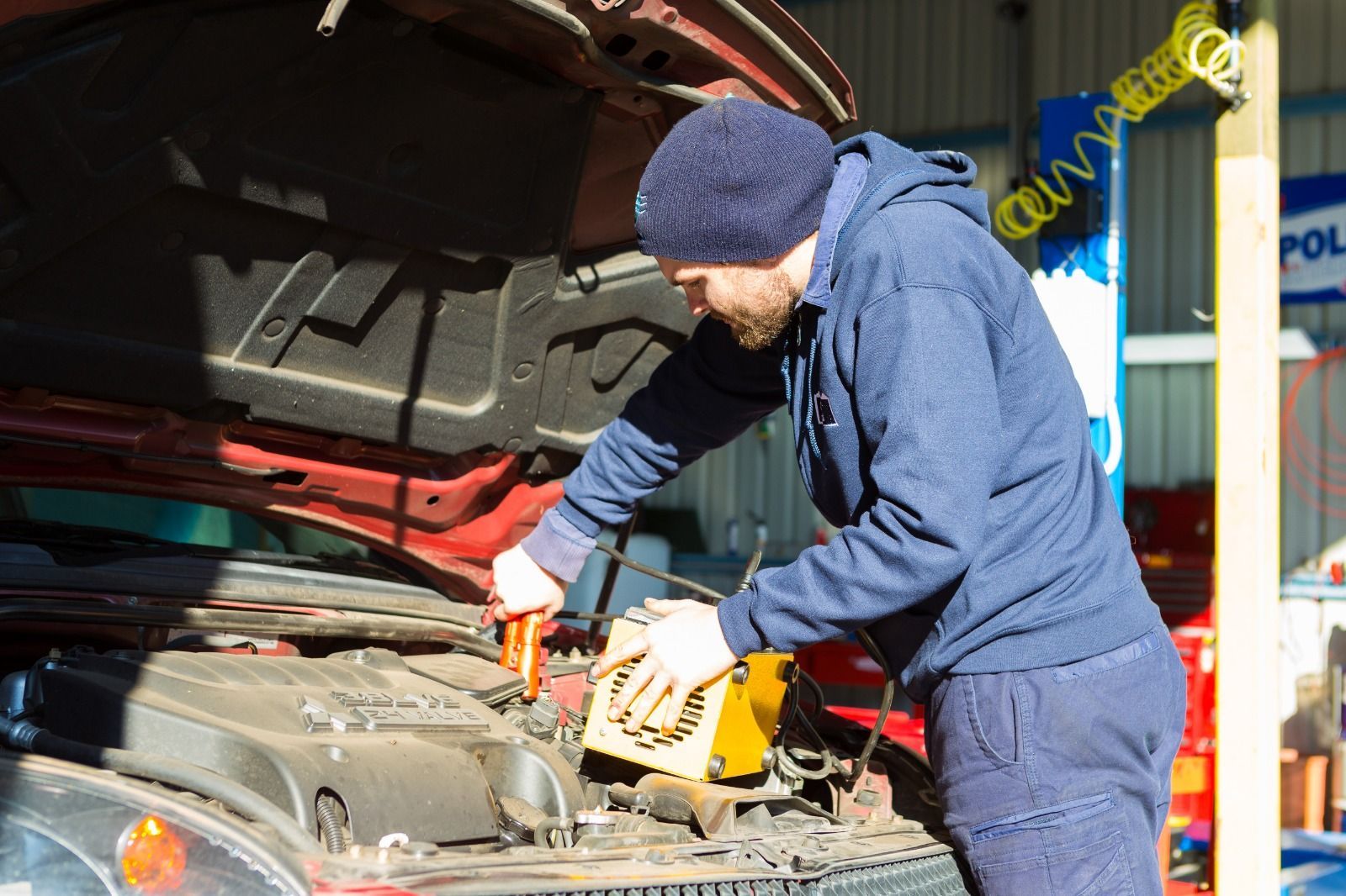 Mechanic in blue coveralls using a jump starter on a red car in a garage.