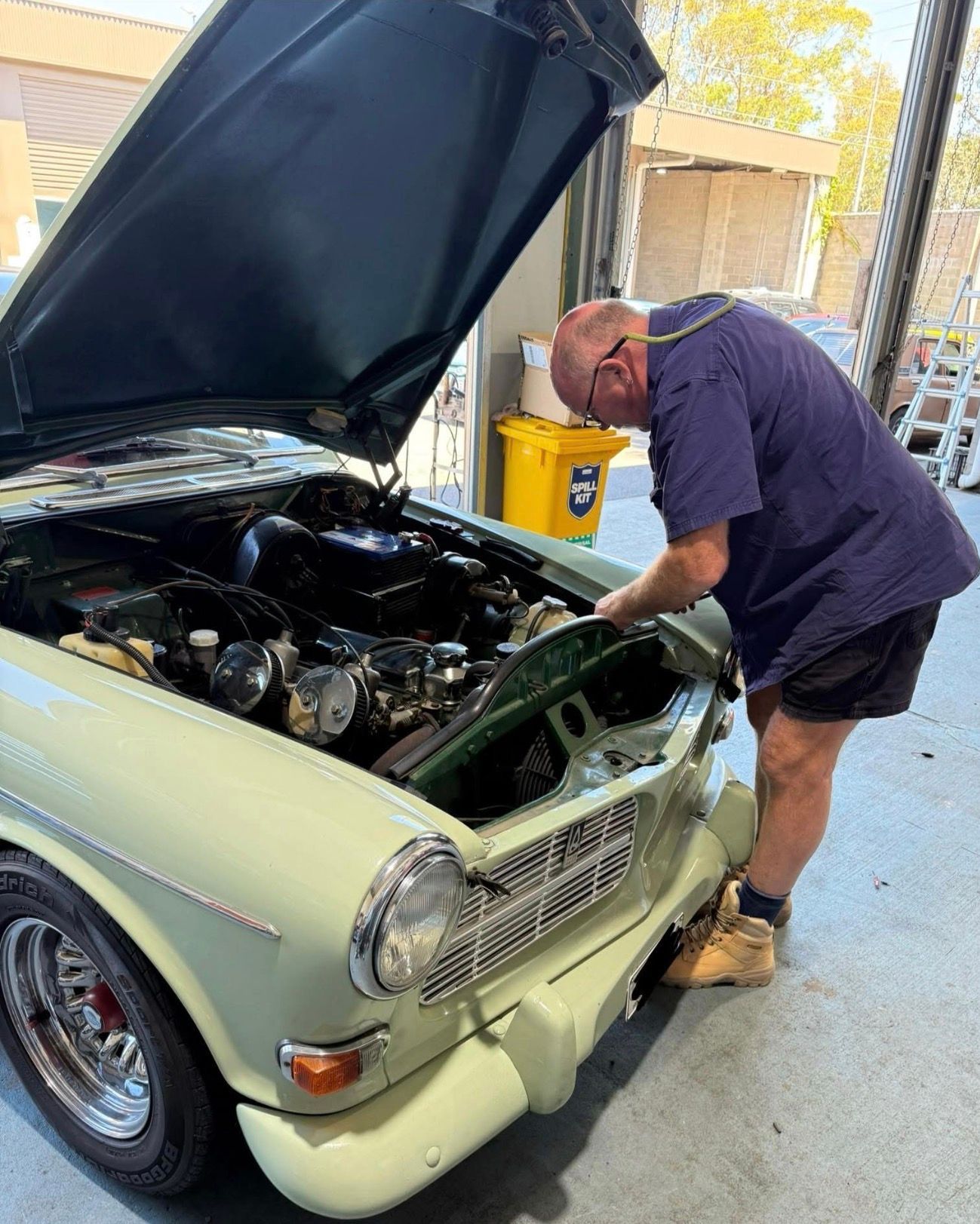 A person in a blue shirt works on the engine of a light green vintage Volvo car with its hood open in a garage.