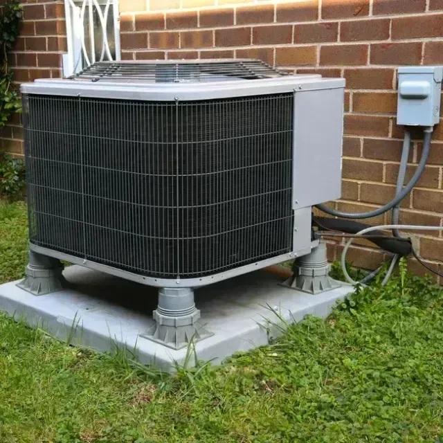 An air conditioning unit on a concrete pad, on a grassy lawn, beside a brick wall.