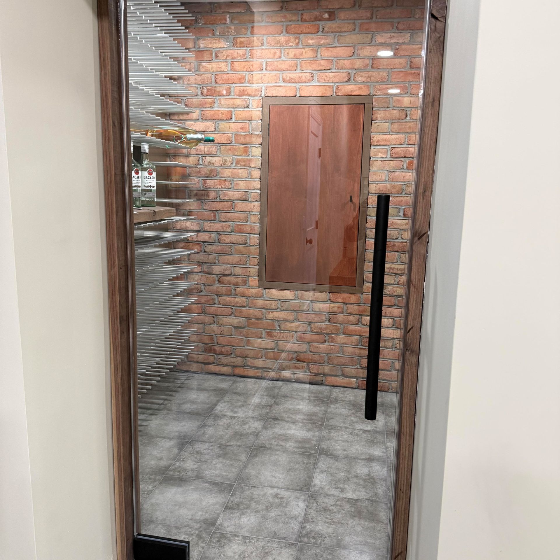 A view through a wooden-framed glass door into a wine cellar with a brick wall, bottle racks, and grey tiled flooring.