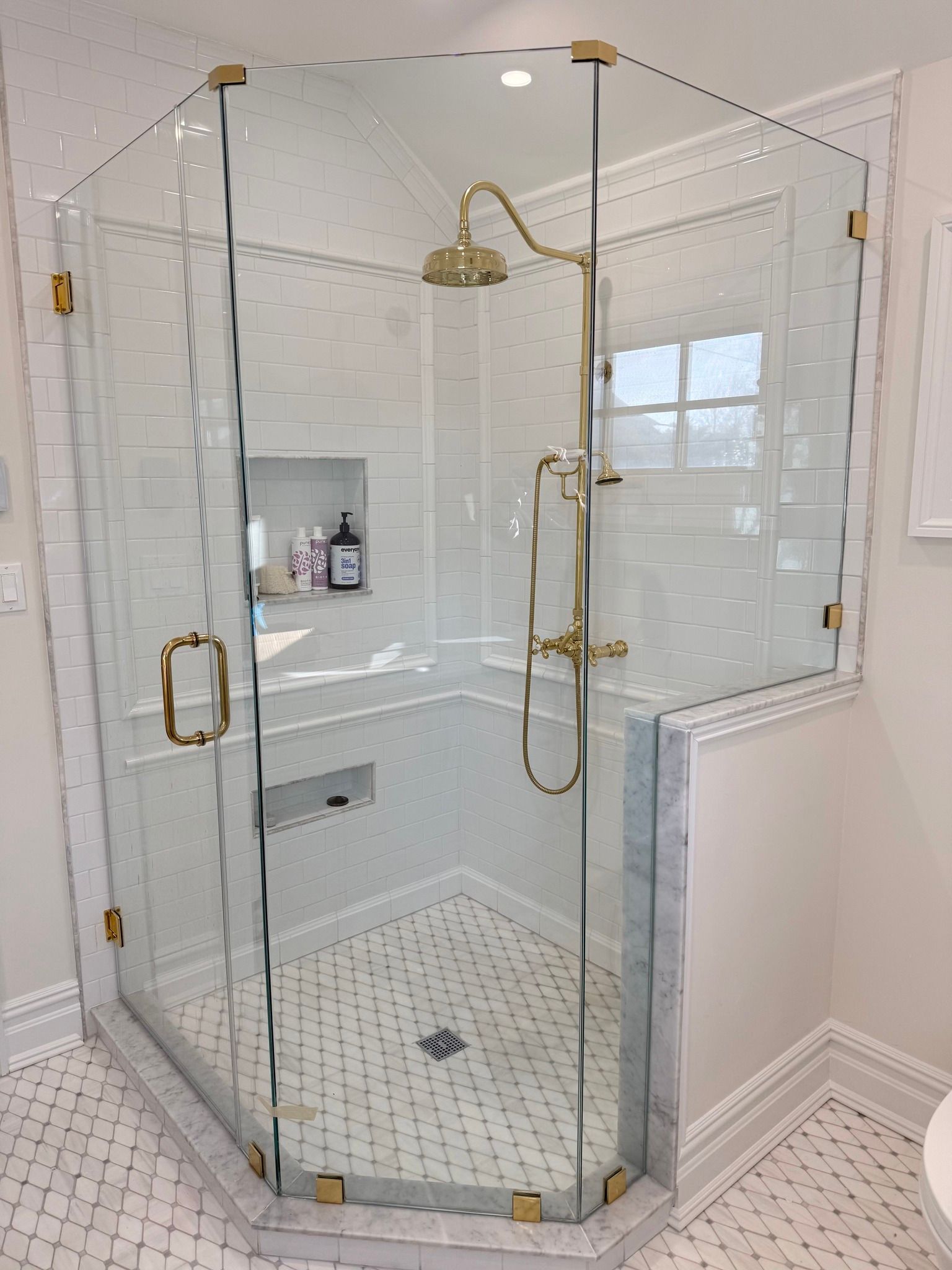 A corner glass shower stall with gold hardware, white tile walls, and a marble patterned floor.
