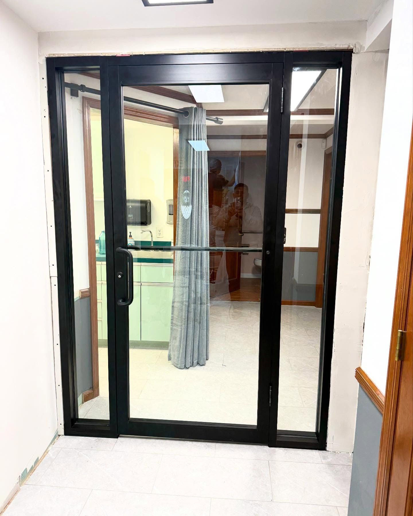 A black-framed glass door with side panels, opening into a bright interior room with a curtain.