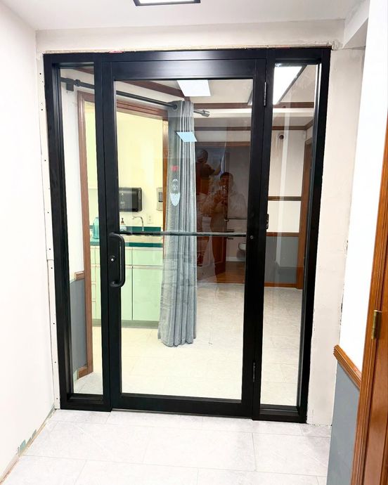 A black metal-framed glass door with side panels, opening into an interior room featuring a patient privacy curtain.