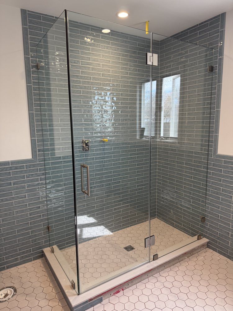 A glass shower enclosure with blue-grey subway tile walls and white hexagonal floor tile.