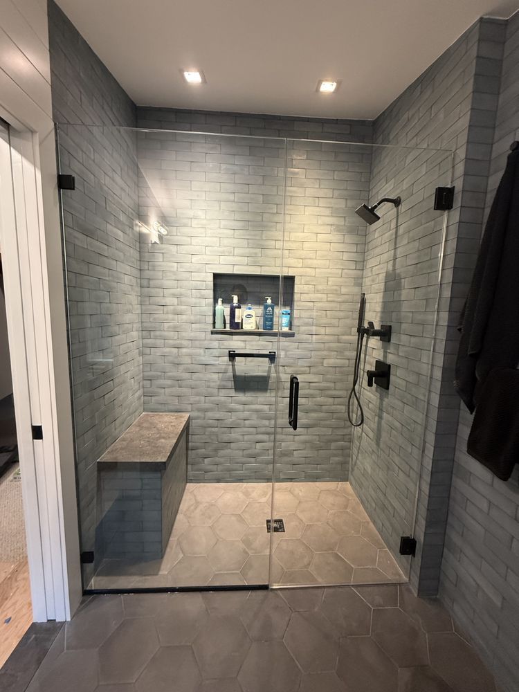A modern walk-in shower with grey brick tile walls, a built-in bench, glass door, and dark fixtures.