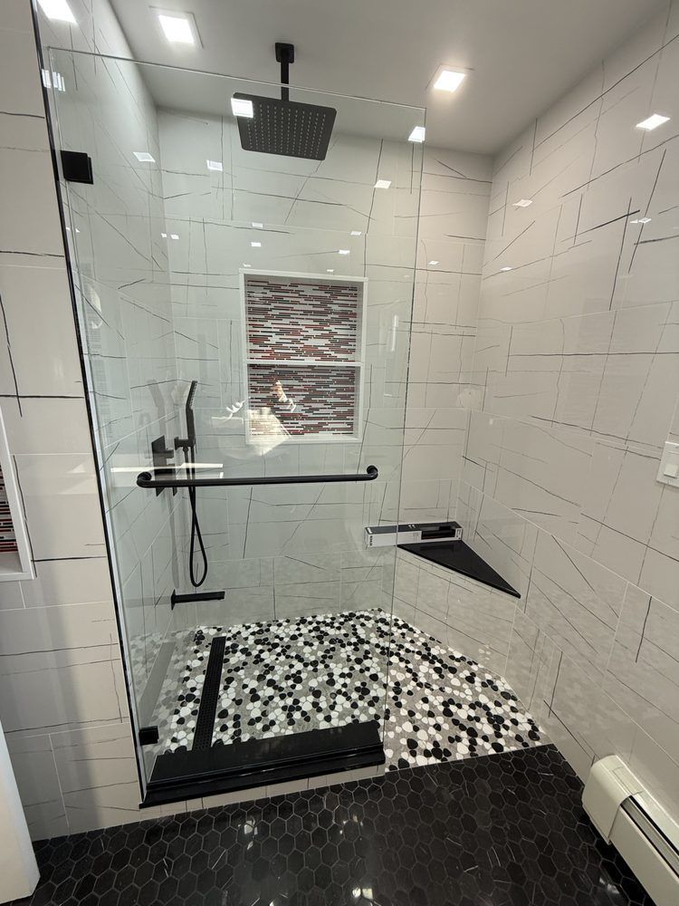 A modern glass walk-in shower with black fixtures, pebble-stone flooring, a corner bench, and white marble-patterned walls.