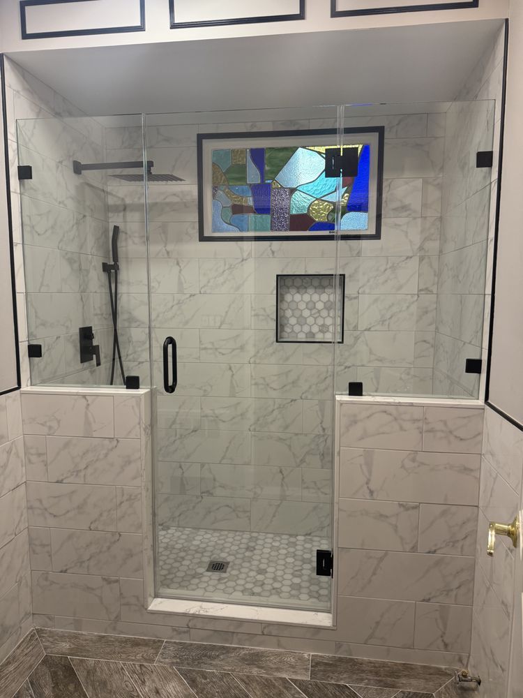 A modern walk-in shower with white marble-patterned tiles, a glass door, black fixtures, and a stained-glass window.