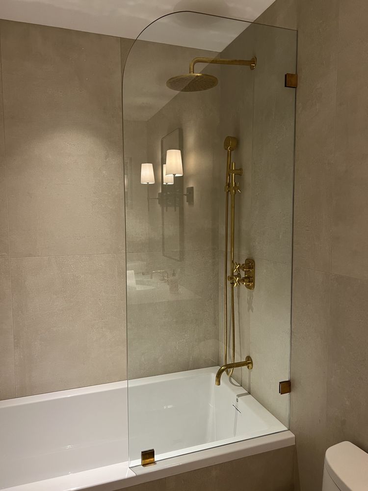 A glass bathtub shower screen with gold fixtures and a rainfall showerhead, mounted in a modern tiled bathroom.