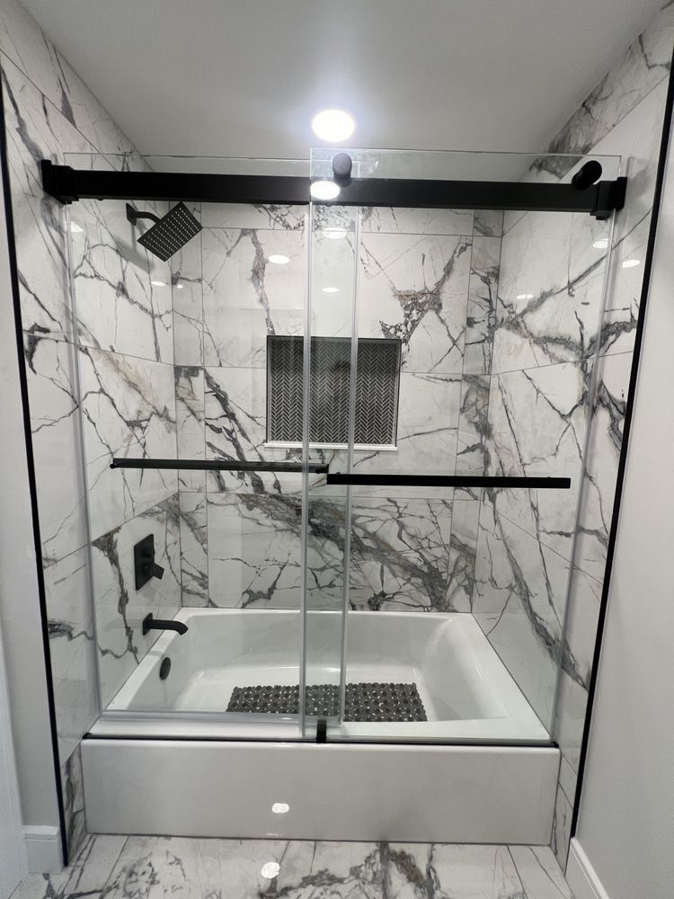 A modern bathroom bathtub and shower enclosure with white marble-patterned tiles and black hardware.