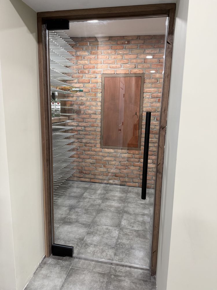 A frameless glass wine cellar door with a wooden frame, leading into a room with a brick wall, shelving, and tile floor.