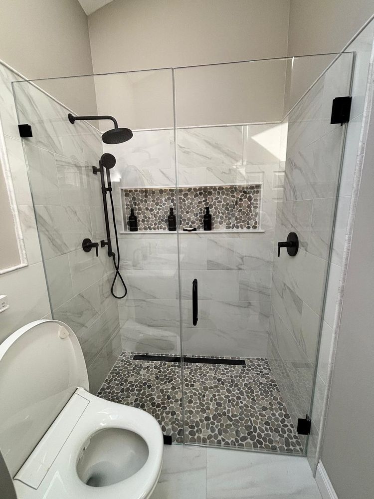 A walk-in shower with white marble-style walls, pebble tile flooring, a horizontal recessed shelf, and black fixtures.