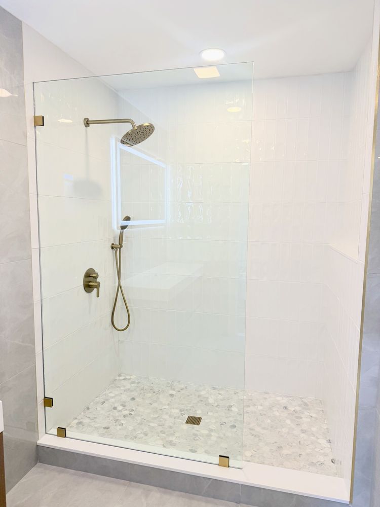 A glass-enclosed shower with white tiled walls, pebble flooring, and brushed gold hardware.